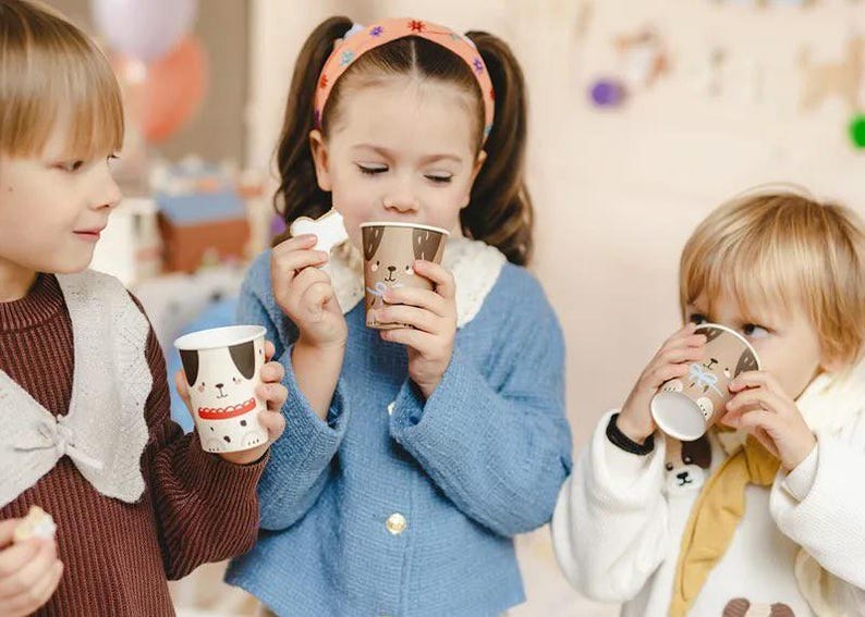 Puede incluir: Tres ni&ntilde;os beben de vasos de papel coloridos con ilustraciones de perros. Los vasos est&aacute;n decorados con la cara de un perro marr&oacute;n y blanco y una nariz roja. Los ni&ntilde;os llevan ropa informal y sonr&iacute;en.