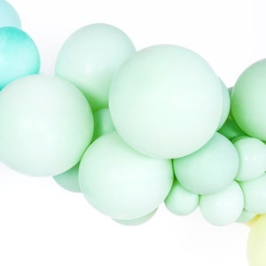 Puede incluir: Una guirnalda de globos de color verde pastel y azul, dispuestos en degradado. Los globos están agrupados, creando un aspecto festivo y caprichoso.