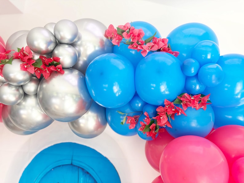 May include: A festive balloon arch featuring silver, blue, and pink balloons, adorned with red flowers. The balloons are arranged in a curved shape, creating a celebratory and decorative display, perfect for parties and events.