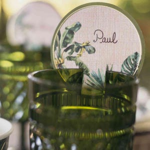 Puede incluir: Un vaso verde con un diseño texturizado, que sostiene una tarjeta de lugar redonda. La tarjeta de lugar tiene un fondo beige claro con un borde verde y un diseño de hojas tropicales. El nombre "Paul" está escrito en cursiva.