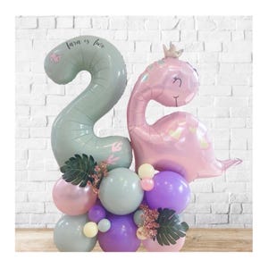 Op de afbeelding: Een feestelijke ballonnenopstelling voor een tweede verjaardag. Met een grote nummer 2 ballon in saliegroen met de tekst "Tara is twee", en een roze dinosaurusvormige ballon. De basis bevat pastelkleurige ballonnen in roze, paars en groen.