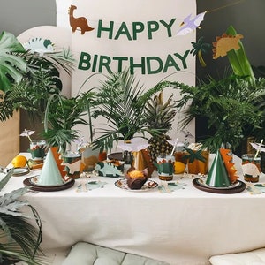 May include: A table setting for a dinosaur-themed birthday party. The table is covered in a white tablecloth and decorated with green and brown dinosaur-shaped plates, cups, and napkins. There is a green dinosaur party hat on the table, as well as a banner that reads "Happy Birthday".