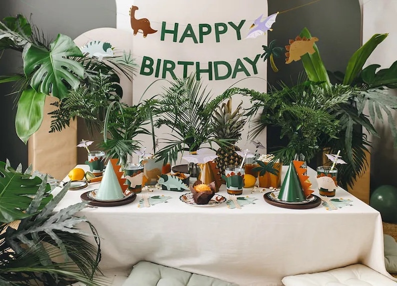 May include: A table setting for a dinosaur-themed birthday party. The table is covered in a white tablecloth and decorated with green and brown dinosaur-shaped plates, cups, and napkins. There is a green dinosaur party hat on the table, as well as a banner that reads "Happy Birthday".