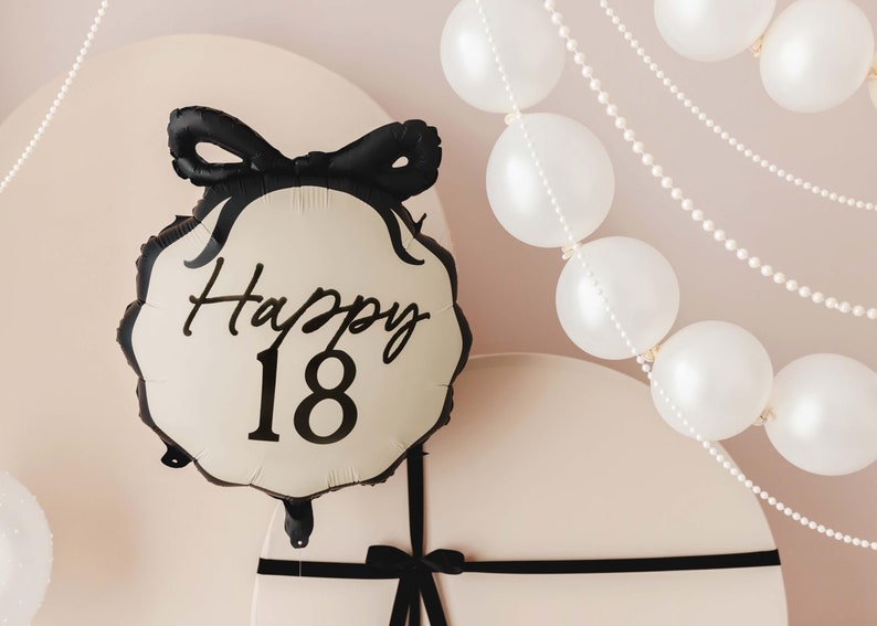 K&ouml;nnte beinhalten: Ein wei&szlig;er Ballon mit schwarzer Aufschrift "Happy 18" und einer schwarzen Schleife. Der Ballon h&auml;ngt an einer wei&szlig;en Decke mit einer Schnur aus wei&szlig;en Perlen.