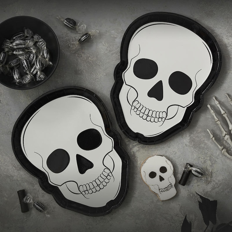 K&ouml;nnte beinhalten: Zwei sch&auml;delformige Pappteller mit schwarzen R&auml;ndern und wei&szlig;en Totenkopfmotiven. Eine kleine Sch&uuml;ssel mit verpackten S&uuml;&szlig;igkeiten, ein sch&auml;delformiger Keks und eine Skeletthand sind ebenfalls vorhanden. Die Artikel sind auf einer grauen Oberfl&auml;che angeordnet, perfekt f&uuml;r Halloween.