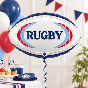 May include: An oval-shaped foil balloon featuring a white textured surface with the word RUGBY printed in large dark blue letters. The design includes red and light blue curved accents, a dark blue border, and a white curling ribbon attached.