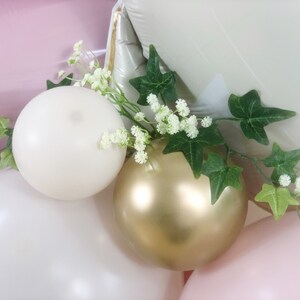 May include: A cluster of balloons in shades of pink, white, and gold, decorated with a sprig of green ivy and white flowers.