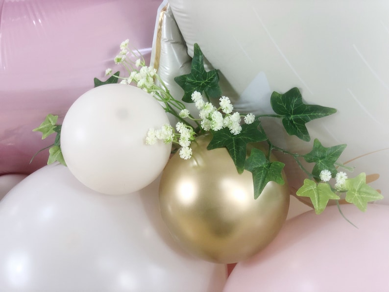 May include: A cluster of balloons in shades of pink, white, and gold, decorated with a sprig of green ivy and white flowers.