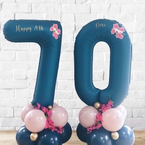 May include: A navy blue number 70 balloon display for a 70th birthday celebration. The balloons are decorated with pink flowers and smaller pink and white balloons at the base. The number 7 balloon has the text "Happy 70th".