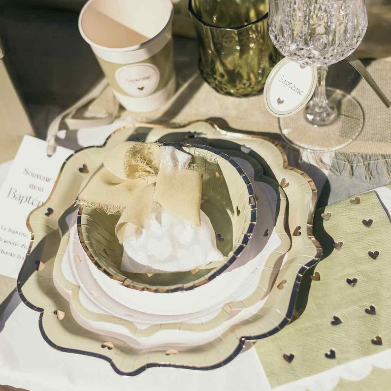 May include: A collection of party supplies, including plates, cups, and a wine glass. The plates are scalloped with gold trim and heart accents. A small gift bag with a ribbon sits in the center. The color palette is soft green and white.