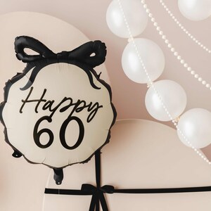 K&ouml;nnte beinhalten: Ein cremefarbener Ballon mit einem schwarzen Band und dem Text "Happy 60" in schwarzer Schrift. Der Ballon ist an einer wei&szlig;en Geschenkbox mit einem schwarzen Band befestigt.