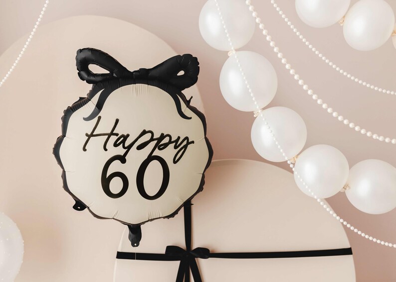 K&ouml;nnte beinhalten: Ein cremefarbener Ballon mit einem schwarzen Band und dem Text "Happy 60" in schwarzer Schrift. Der Ballon ist an einer wei&szlig;en Geschenkbox mit einem schwarzen Band befestigt.