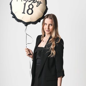 K&ouml;nnte beinhalten: Eine Frau in einem schwarzen Blazer h&auml;lt einen goldenen Ballon mit schwarzem Besatz und dem aufgedruckten Text "Happy 18".