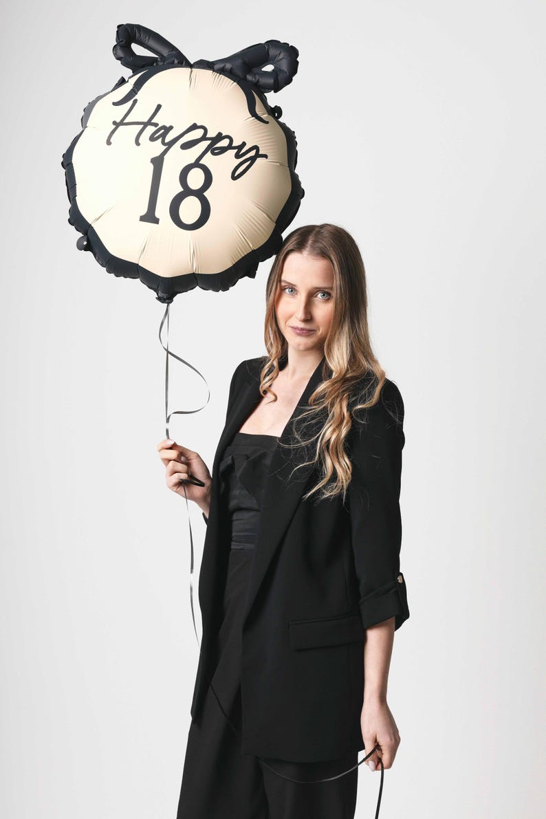 K&ouml;nnte beinhalten: Eine Frau in einem schwarzen Blazer h&auml;lt einen goldenen Ballon mit schwarzem Besatz und dem aufgedruckten Text "Happy 18".