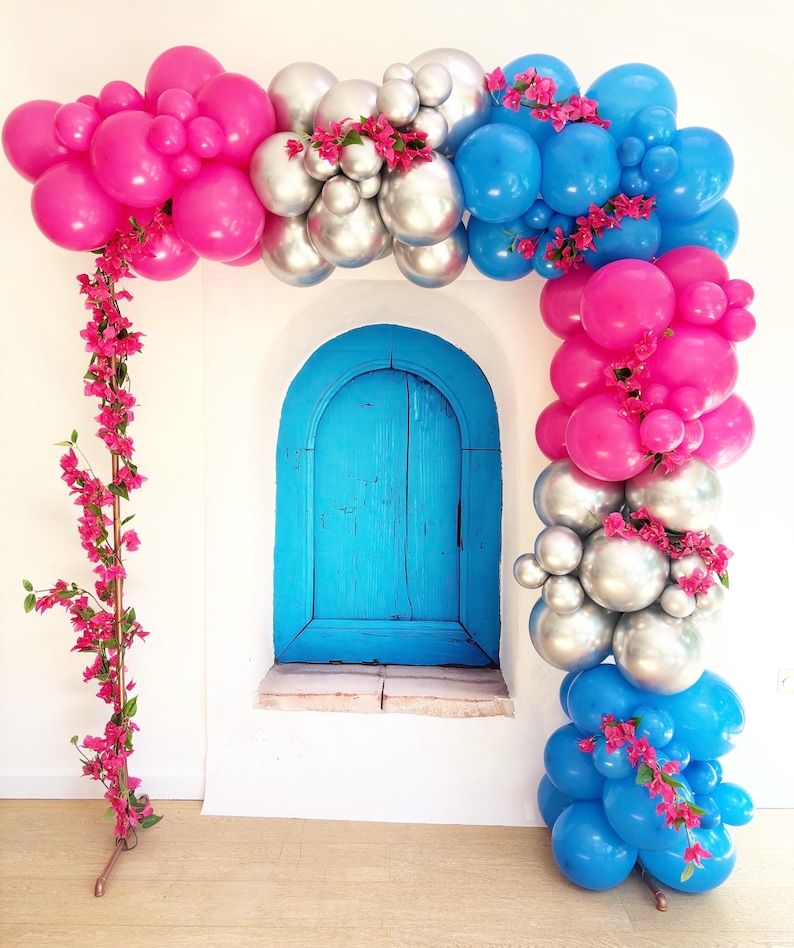 May include: A vibrant balloon arch in shades of pink, silver, and blue, adorned with pink floral accents. The arch frames a bright blue arched doorway set against a white wall. A decorative piece for parties and celebrations.