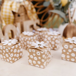 Op de afbeelding: Verschillende kleine, vierkante geschenkdozen met een madeliefjespatroon op een lichtbruine achtergrond. Elke doos is bekroond met een witte madelief. De dozen zijn gerangschikt op een lichtgekleurd oppervlak, met vage decoratieve elementen op de achtergrond.