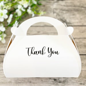 Può includere: Scatola regalo bianca con un manico a forma di borsa. La scatola ha le parole "Grazie" stampate sul davanti.