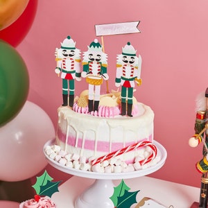 Op de afbeelding: Een witte en roze taart versierd met roze en witte frosting en bedekt met notenkrakerfiguurtjes. De taart is versierd met een snoepstok en witte marshmallows. De taart staat op een witte taartstandaard met een witte achtergrond.