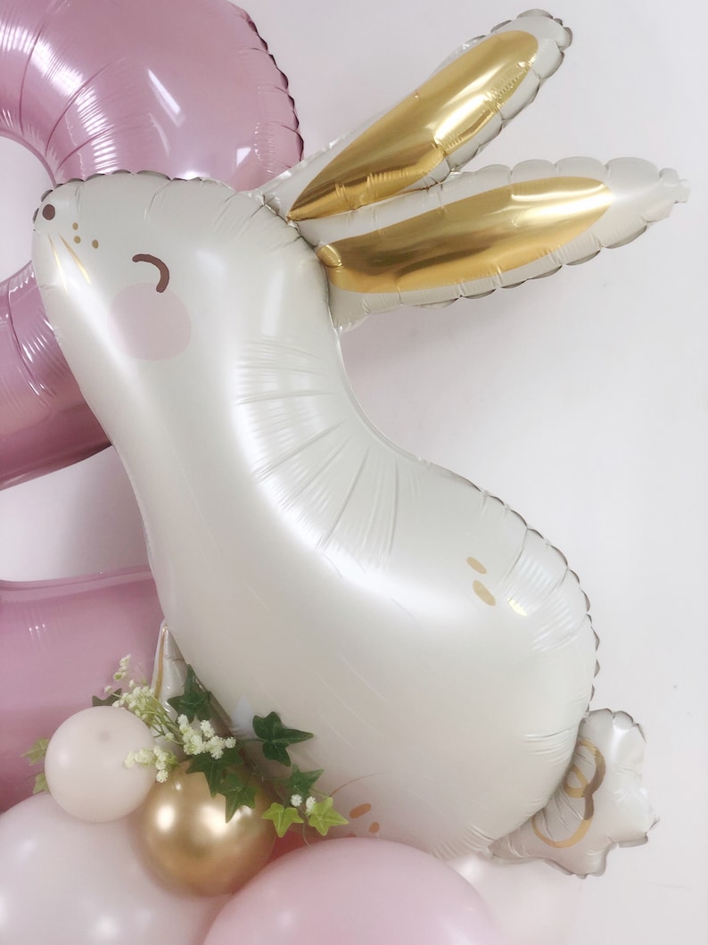 May include: A white balloon shaped like a bunny with gold ears and a pink nose. The bunny is surrounded by pink, white, and gold balloons.