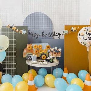 Pode incluir: Um conjunto de decora&ccedil;&atilde;o de festa de anivers&aacute;rio com tema de constru&ccedil;&atilde;o com um grande bal&atilde;o de 'Feliz anivers&aacute;rio', uma faixa com caminh&otilde;es de constru&ccedil;&atilde;o e uma variedade de bal&otilde;es amarelos, azuis e verdes. O conjunto tamb&eacute;m inclui um caminh&atilde;o basculante de brinquedo, uma mesa com donuts e cupcakes, e uma variedade de outras decora&ccedil;&otilde;es.
