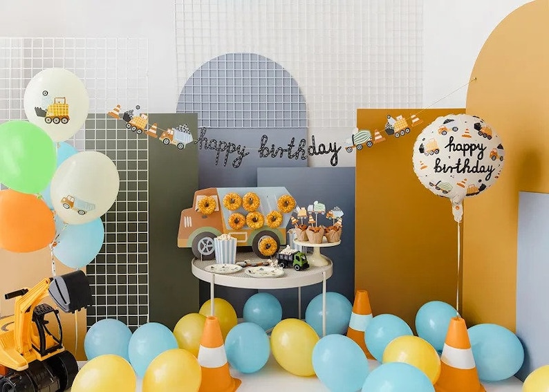 Pode incluir: Um conjunto de decora&ccedil;&atilde;o de festa de anivers&aacute;rio com tema de constru&ccedil;&atilde;o com um grande bal&atilde;o de 'Feliz anivers&aacute;rio', uma faixa com caminh&otilde;es de constru&ccedil;&atilde;o e uma variedade de bal&otilde;es amarelos, azuis e verdes. O conjunto tamb&eacute;m inclui um caminh&atilde;o basculante de brinquedo, uma mesa com donuts e cupcakes, e uma variedade de outras decora&ccedil;&otilde;es.