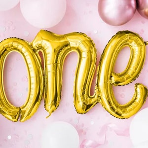 Può includere: Palloncini in lamina dorata che compongono la parola "uno" su uno sfondo rosa con coriandoli bianchi e rosa.