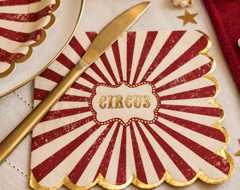 May include: A square paper napkin with a red and white sunburst pattern and a scalloped gold foil edge. The center features a decorative white label with the word CIRCUS printed in gold lettering. A gold-toned metal knife rests on top.