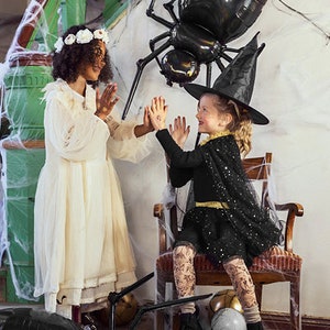 May include: Two children dressed in Halloween costumes. One child is wearing a white dress with a flower crown and the other is wearing a black witch costume with a black hat. Both children are standing in front of a large black spider balloon.