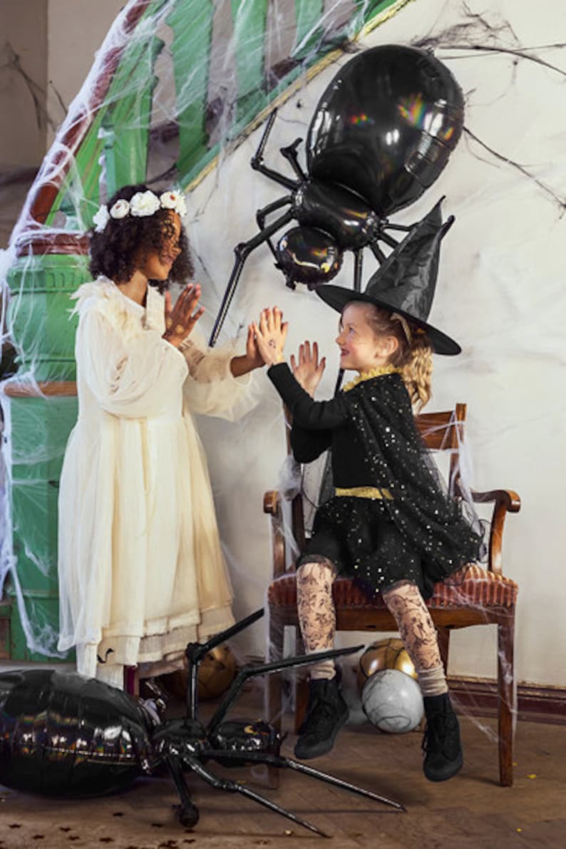 May include: Two children dressed in Halloween costumes. One child is wearing a white dress with a flower crown and the other is wearing a black witch costume with a black hat. Both children are standing in front of a large black spider balloon.