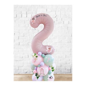 Puede incluir: Un globo rosa con el número 2 y el texto "Tara is two" es el centro de un arreglo de globos. El arreglo incluye globos pastel más pequeños, pegatinas de hadas y vegetación, creando una decoración de cumpleaños caprichosa.