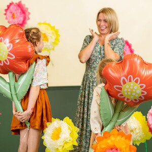 May include: Two children hold large red flower-shaped balloons with green stems. A woman in a floral dress smiles at the children. The background is decorated with paper flowers in various colors.