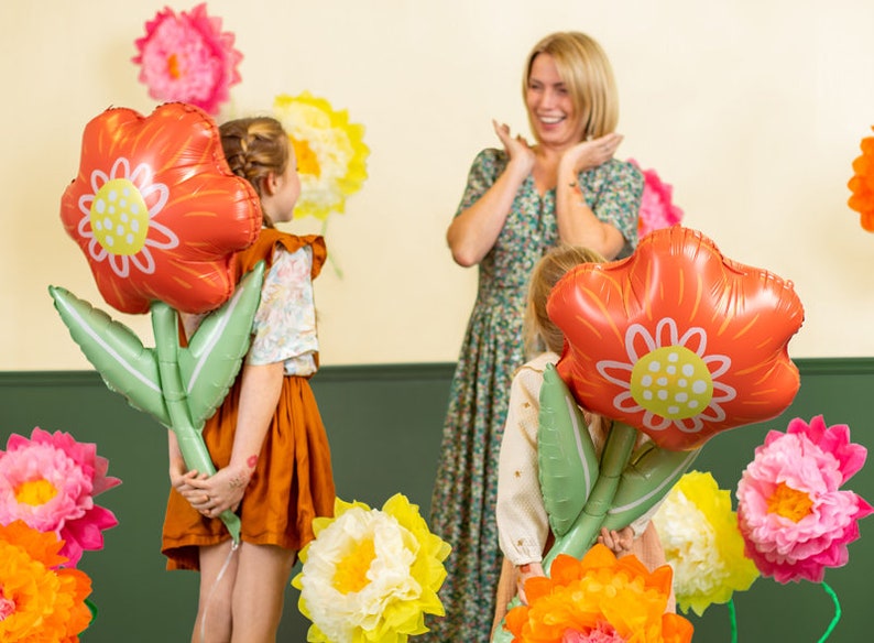 May include: Two children hold large red flower-shaped balloons with green stems. A woman in a floral dress smiles at the children. The background is decorated with paper flowers in various colors.