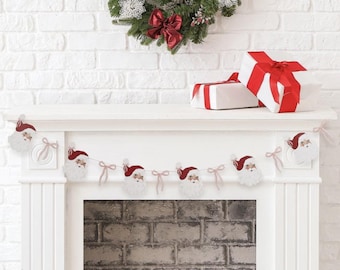 Puede incluir: Una repisa de chimenea navideña decorada con una corona festiva, regalos y una guirnalda de Papá Noel. La corona presenta lazos rojos y adornos. Los regalos están envueltos en papel blanco con cintas rojas. La guirnalda tiene caras de Papá Noel.