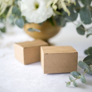 Op de afbeelding: Twee kleine, vierkante, bruine kartonnen dozen staan op een witte ondergrond, met een vage bloemstuk op de achtergrond. De dozen zijn effen, wat suggereert dat ze voor het verpakken van kleine items zijn.