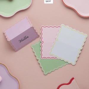 May include: A selection of pastel place cards with a scalloped design. One card is lavender with the name "Millie" in script. Other cards are pink, green, and white, each with a contrasting border. The cards are arranged on a pink surface.