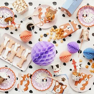 Puede incluir: Una escena festiva con decoraciones tem&aacute;ticas de perros. Incluye platos, vasos y cajas con ilustraciones de corgis, galletas con forma de hueso, palomitas de ma&iacute;z y bolas de papel de nido de abeja en varios colores. El mantel tiene un estampado de lunares blancos y negros.