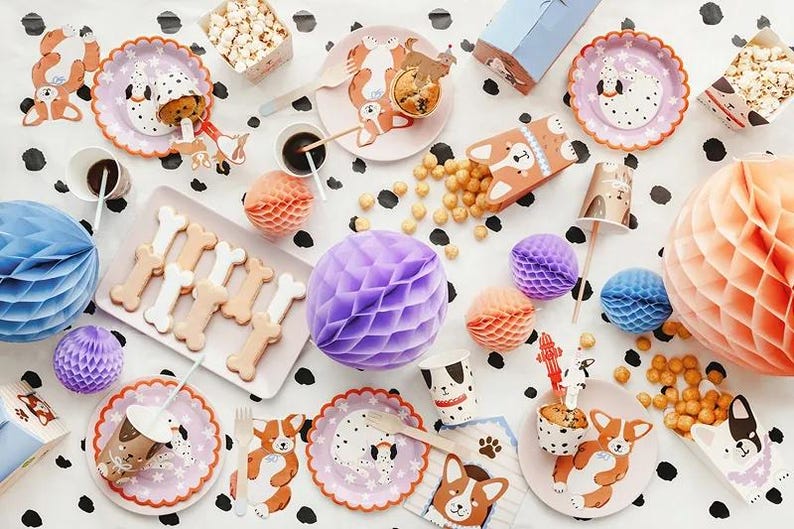 Puede incluir: Una escena festiva con decoraciones tem&aacute;ticas de perros. Incluye platos, vasos y cajas con ilustraciones de corgis, galletas con forma de hueso, palomitas de ma&iacute;z y bolas de papel de nido de abeja en varios colores. El mantel tiene un estampado de lunares blancos y negros.