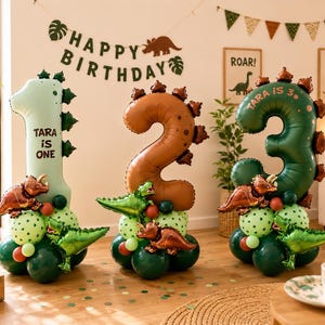 May include: Three large number-shaped foil balloons representing one, two, and three. The light green one says 'TARA IS ONE', the brown two has dinosaur spikes, and the dark green three says 'TARA IS 3'. Each balloon sits on a base of smaller balloons.