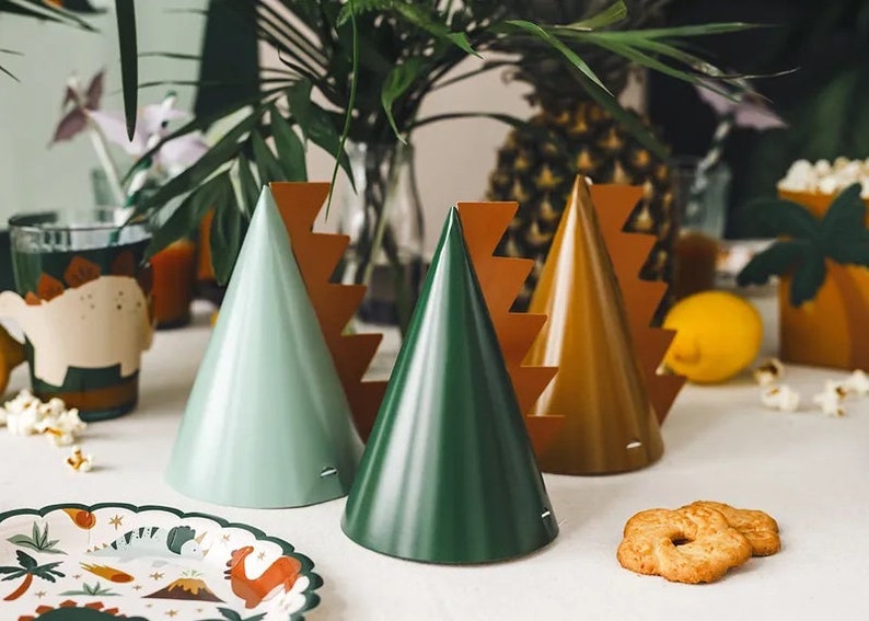 May include: A set of three party hats in green, light green, and brown with a zig-zag pattern around the base. The hats are on a white table with a plate and cookies.