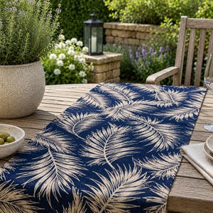 May include: A navy blue table runner with a repeating pattern of beige palm leaves. The runner is placed on a wooden table, alongside a potted plant, a bowl of olives, and wine glasses. The scene is set outdoors.