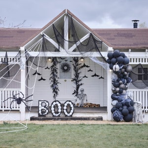 May include: A white house decorated for Halloween with a large spider web, a giant spider, and the word "BOO" spelled out with white spheres. Black balloons, bats, and a skeleton complete the spooky scene.