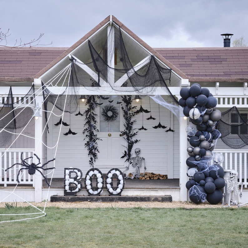 May include: A white house decorated for Halloween with a large spider web, a giant spider, and the word "BOO" spelled out with white spheres. Black balloons, bats, and a skeleton complete the spooky scene.