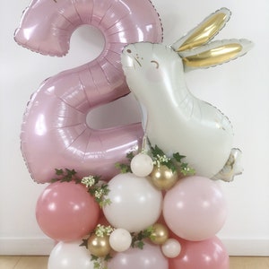 May include: A pink number two balloon with the text "Tara is Two" on it, a white bunny balloon, and a cluster of pink, white, and gold balloons with greenery.