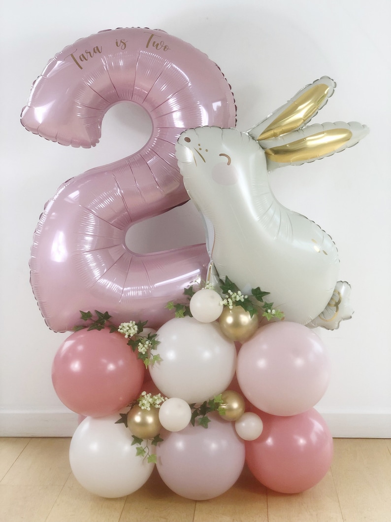 May include: A pink number two balloon with the text "Tara is Two" on it, a white bunny balloon, and a cluster of pink, white, and gold balloons with greenery.