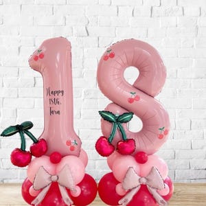 May include: Pink number balloons, shaped as the numbers 1 and 8, decorated with cherry designs. The number 1 has the text "Happy 18th. Tara". The balloons are on a base of pink and red balloons with bows and cherry accents.
