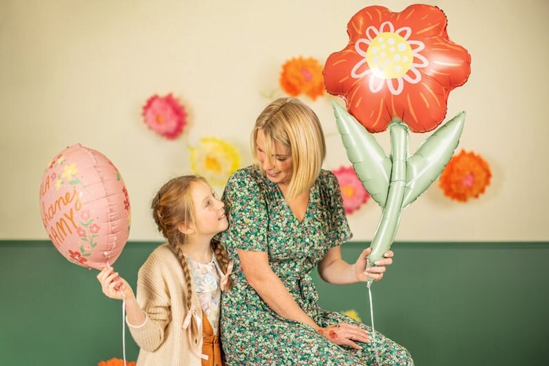 May include: A mother and daughter are holding pink and red foil balloons shaped like flowers. The balloons have floral designs and the pink balloon says "Happy Mama".