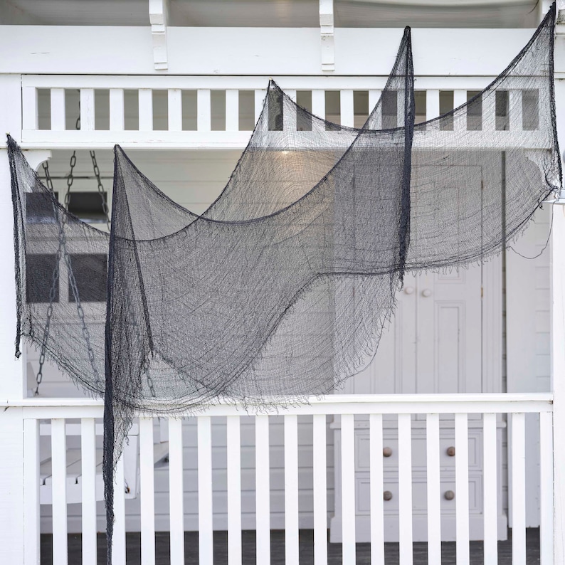 May include: Black, tattered fabric draped over a white porch railing and ceiling. The fabric is hung in a wavy pattern, creating a spooky, Halloween-themed decoration. The porch has a white railing and a white door.