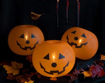 May include: Three orange pumpkin-shaped decorations with black jack-o'-lantern faces are lit from within. Black candles with red wax drip are in the background, along with autumn leaves, creating a Halloween theme.