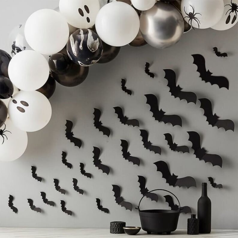 May include: Black and white balloons with ghost faces and a silver balloon are arranged in an arch. Black paper bats are scattered on a gray wall. A black cauldron and other Halloween decorations are on a table in the foreground.