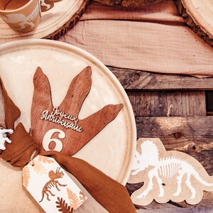 May include: Dinosaur-themed birthday decorations. A beige plate features a brown dinosaur footprint, the number 6, and the words "Joyeux Anniversaire". A tag with a dinosaur illustration is attached.